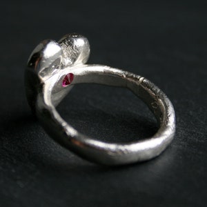 May include: A silver ring with a textured band and a heart-shaped design. A small red gemstone is set in the center of the heart.