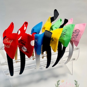 May include: A set of six colorful fabric headbands with bows. The bows are decorated with various Disney characters, including Mickey Mouse, Minnie Mouse, and a fairy. The headbands are displayed on a clear acrylic stand.