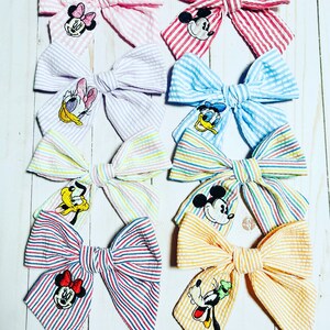 May include: Eight striped fabric hair bows with different Disney character embroidery. The bows are in pink, purple, blue, yellow, red, and orange.