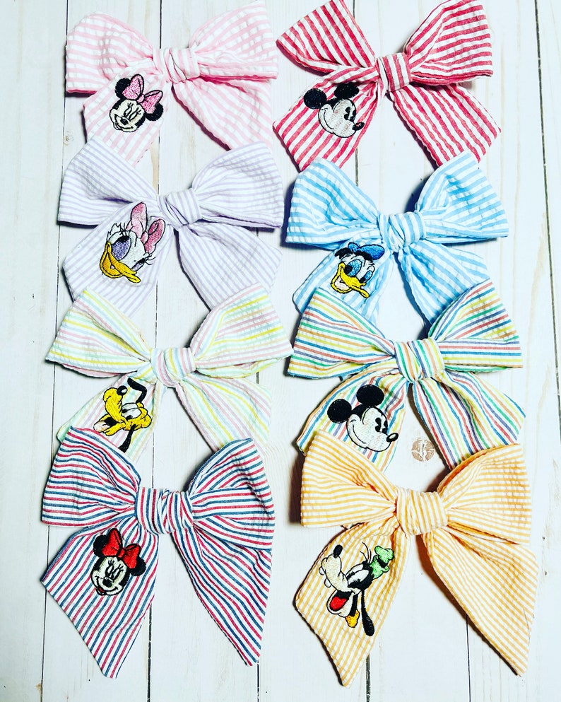 May include: Eight striped fabric hair bows with different Disney character embroidery. The bows are in pink, purple, blue, yellow, red, and orange.