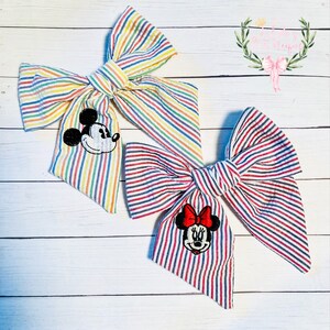May include: Two fabric hair bows with striped patterns. One bow is rainbow striped with a Mickey Mouse applique. The other bow is red and white striped with a Minnie Mouse applique.