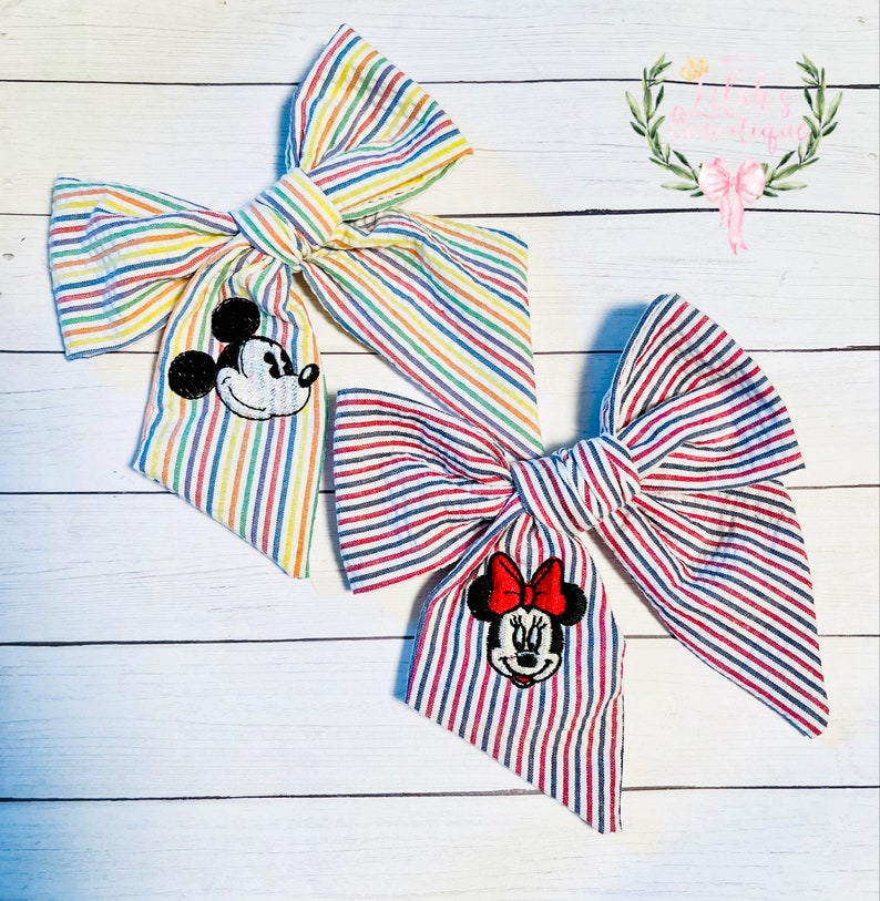 May include: Two fabric hair bows with striped patterns. One bow is rainbow striped with a Mickey Mouse applique. The other bow is red and white striped with a Minnie Mouse applique.