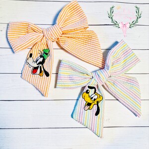 May include: Two striped fabric hair bows with embroidered cartoon dog characters. The bow on the left is orange and white with a Goofy character. The bow on the right is rainbow striped with a Pluto character.