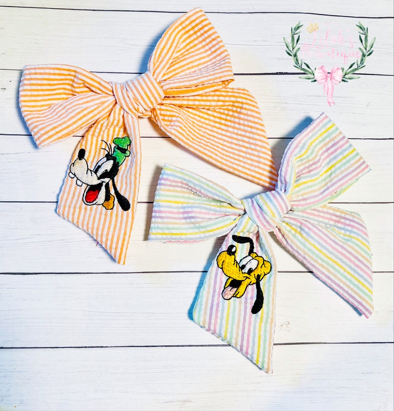 May include: Two striped fabric hair bows with embroidered cartoon dog characters. The bow on the left is orange and white with a Goofy character. The bow on the right is rainbow striped with a Pluto character.