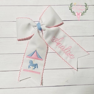 May include: White bow with pink trim and a blue ribbon. The bow has an embroidered carousel horse and the name "Adelaide" on it.
