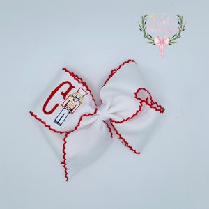 May include: White bow with red scalloped edges and a red and blue embroidered nutcracker and the letter "C".