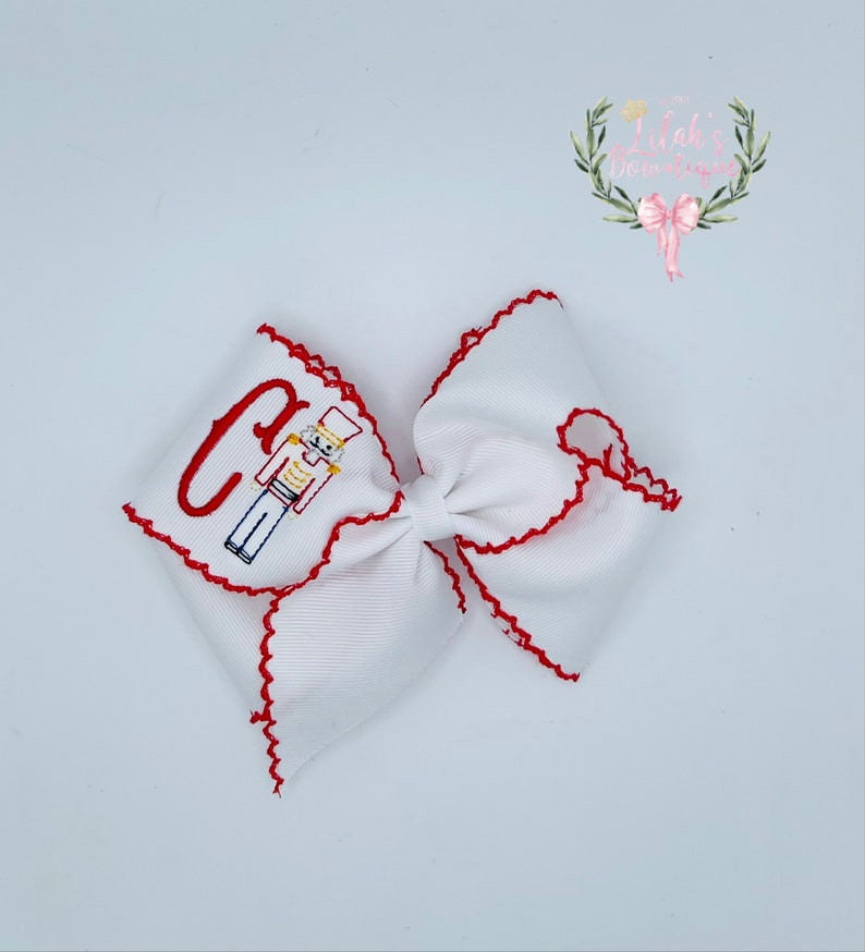 May include: White bow with red scalloped edges and a red and blue embroidered nutcracker and the letter "C".