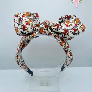 May include: A white headband with a bow featuring a repeated pattern of Chip 'n Dale cartoon characters. The headband is on a clear display stand.