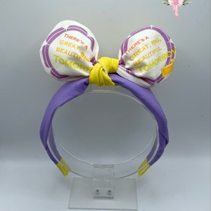 May include: Purple headband with a yellow bow and white fabric ears. The ears have the text "There's a Great, Big, Beautiful Tomorrow" printed on them.