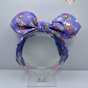 May include: A purple headband with a large bow featuring a Daisy Duck print. The bow and headband are decorated with white polka dots and daisy flowers. The headband has pink accents on the ends.