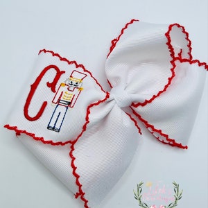 May include: White hair bow with red scalloped edges and an embroidered nutcracker design with the letter "C".