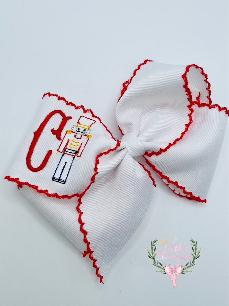 May include: White hair bow with red scalloped edges and an embroidered nutcracker design with the letter "C".