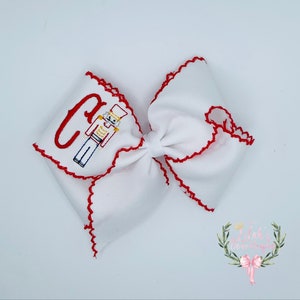 May include: White bow with red scalloped edges and a red and yellow embroidered nutcracker and the letter C.