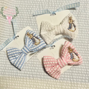 May include: Three seersucker bow ties in blue, white, and pink, each featuring an embroidered Peter Rabbit design. The bows are attached to white cards with blue gingham ribbons for hanging. The background is a textured white fabric.