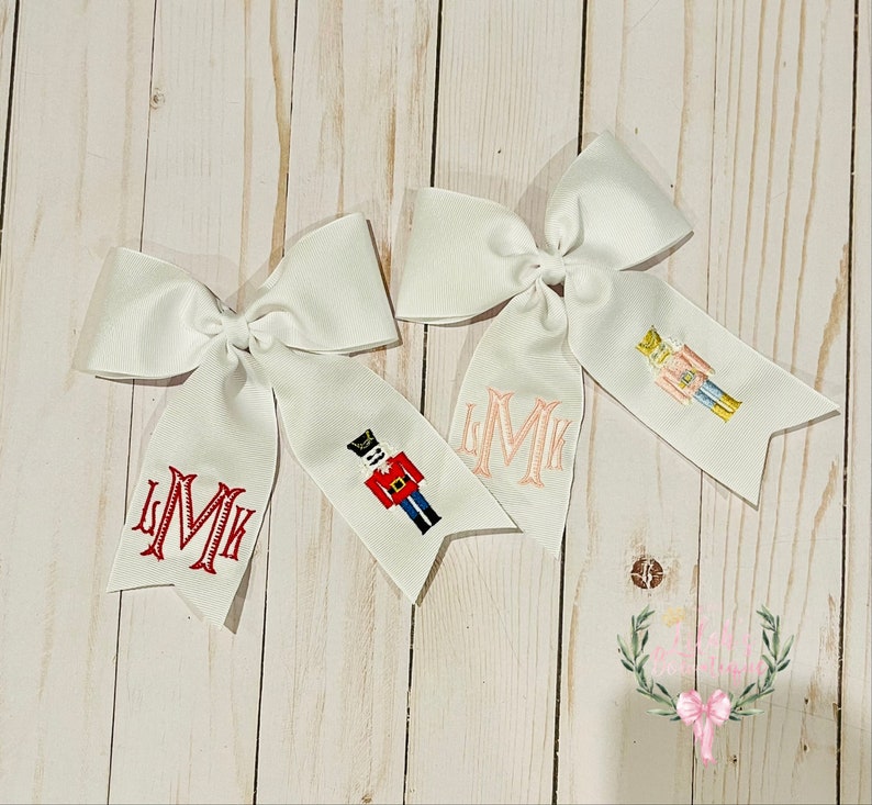K&ouml;nnte beinhalten: Zwei wei&szlig;e Haarb&auml;nder mit einem Nussknacker-Design und dem Monogramm "LMK". Ein Band hat einen rosa Nussknacker und das andere einen roten Nussknacker.