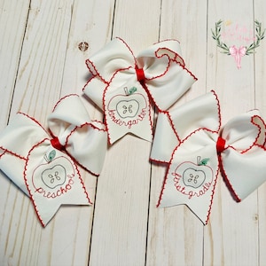 May include: Three white hair bows with red trim and an embroidered apple design. Each bow has a different grade level embroidered on it: "Preschool", "Kindergarten", and "First Grade".