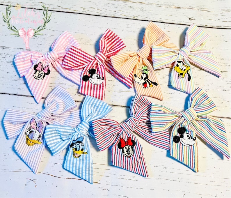 May include: Set of eight striped fabric hair bows with various Disney character embroidery. The bows are in pink, red, blue, orange, and rainbow colors.