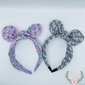 May include: Two fabric headbands with geometric patterns. One headband is pink, blue, and purple. The other headband is gray. Both headbands have a bow in the center and are tied into a knot.
