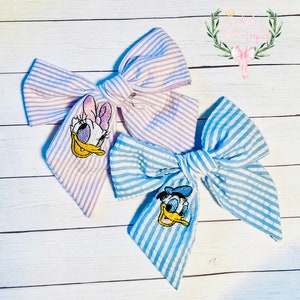 May include: Two fabric hair bows with embroidered cartoon characters. The bow on the left is purple and white striped with a pink and white embroidered Daisy Duck. The bow on the right is blue and white striped with a blue and yellow embroidered Donald Duck.