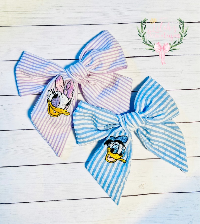 May include: Two fabric hair bows with embroidered cartoon characters. The bow on the left is purple and white striped with a pink and white embroidered Daisy Duck. The bow on the right is blue and white striped with a blue and yellow embroidered Donald Duck.