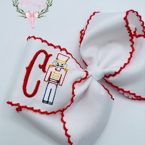 May include: White bow with red scalloped edges and a red embroidered letter C and a nutcracker design.
