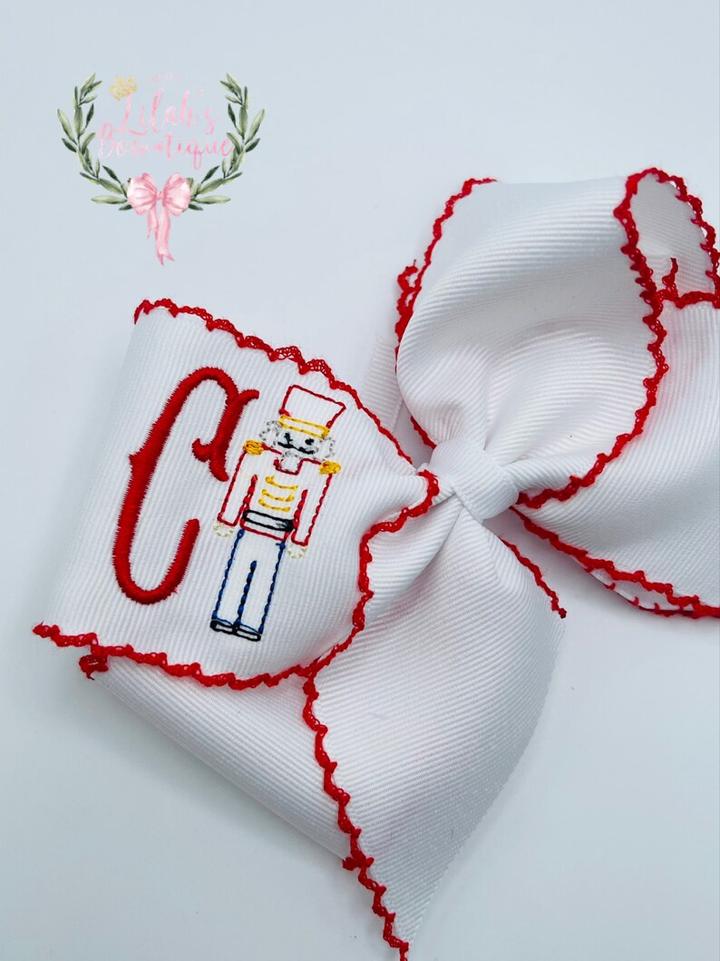 May include: White bow with red scalloped edges and a red embroidered letter C and a nutcracker design.