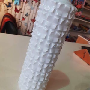 Puede incluir: Un jarrón blanco con un diseño geométrico texturizado. El jarrón tiene forma cilíndrica y una parte superior ensanchada. La superficie está cubierta con un patrón repetitivo de diseños en forma de diamante en relieve. El jarrón mide unos 30 cm de alto.