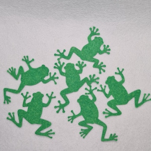 May include: Six green felt frog cutouts are scattered on a white surface. The frogs are in various poses, with outstretched limbs and webbed feet. The felt has a slightly textured appearance, and the frogs are a vibrant shade of green.