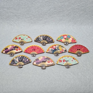 May include: Assortment of ten wooden buttons shaped like fans with various floral patterns in shades of blue, pink, red, and purple. The buttons have a gold trim around the edges.