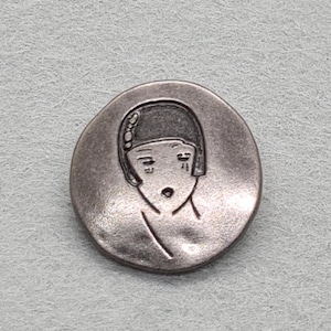 May include: A circular, antique silver-coloured button featuring a stylised Art Deco woman's face. The button has a detailed engraving of a woman's face with a bob haircut and a surprised expression. The button is set against a light grey background.