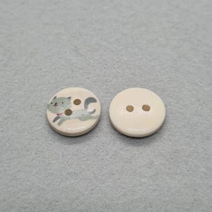 May include: Two round wooden buttons. One button features a gray cat illustration with brown spots and a pink collar. The other button is plain with two holes for sewing. Both buttons are off-white.