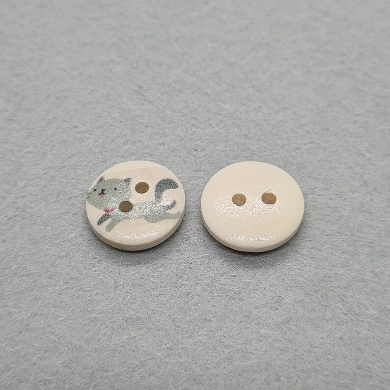 May include: Two round wooden buttons. One button features a gray cat illustration with brown spots and a pink collar. The other button is plain with two holes for sewing. Both buttons are off-white.