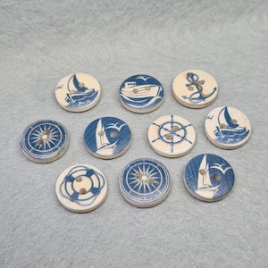 May include: Ten wooden buttons with blue and white nautical designs. The designs include a sailboat, a compass, a life preserver, an anchor, and a ship's wheel.