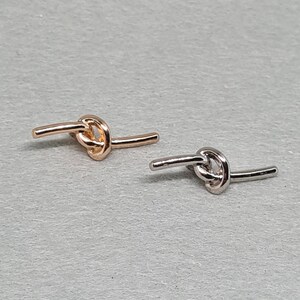May include: Two small, knot-shaped earrings. One is rose gold, the other is silver. The earrings are on a light grey surface. The knot design is a simple, elegant style.