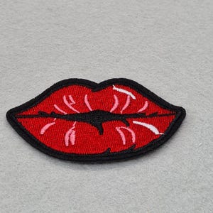 May include: An embroidered patch in the shape of a pair of lips. The lips are bright red with a thick black border and a black centre line. The design features white and light pink stitched highlights that create a sense of depth and texture.