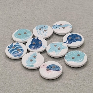 May include: A collection of ten round, white wooden buttons with sea creature designs. Each button features a different marine animal, including a fish, jellyfish, whale, and turtle, in shades of blue and teal. The buttons have two small holes for sewing.