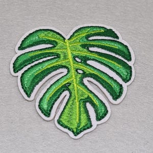 May include: Embroidered patch in the shape of a Monstera leaf. The leaf is a vibrant green with lighter green veins and a white border. The patch is designed to be ironed or sewn onto fabric.