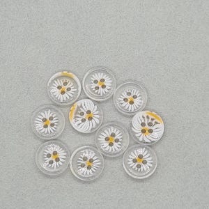 May include: A collection of clear, round buttons with a daisy design. Each button features a white daisy with a yellow center and is set against a clear background. The buttons are scattered on a gray surface.