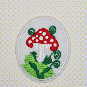 May include: A white felt patch featuring a red and white mushroom with white spots and green leaves. The mushroom is embroidered with white and red thread, and the leaves are embroidered with green thread.
