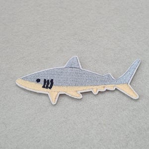 May include: An embroidered iron-on patch shaped like a shark. The shark features a light grey upper body and a pale yellow underbelly, with a dark black eye and three vertical gill slits. The entire design is outlined in a white border.