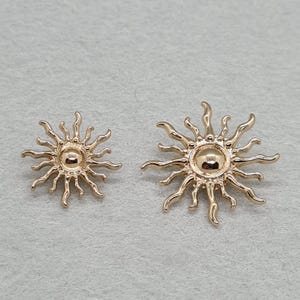 May include: Two gold-toned metal sun-shaped charms with a polished finish. Each charm features a central circular dome surrounded by radiating, wavy sunbeams. The charms are identical in design but differ in size, with one being smaller.