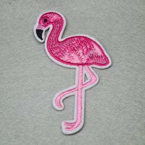May include: Pink flamingo embroidered iron-on patch. The flamingo is standing on one leg with its neck curved. The patch has a white outline.