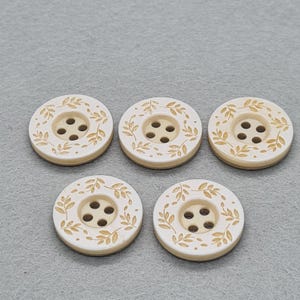 May include: Five round, off-white buttons with a light beige leaf design around the edges. Each button has four holes in the centre for sewing. The buttons are made of a smooth material.