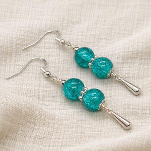 Peut inclure: Une paire de boucles d'oreilles pendantes composées de deux perles rondes en verre craquelé turquoise, suspendues à des crochets en métal argenté. Les boucles se terminent par une petite breloque en forme de goutte en métal argenté lisse.