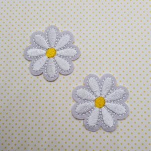 May include: Two white felt daisy flower patches with yellow centres. The daisies are embroidered with white thread and have a grey outline.