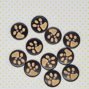 May include: A selection of black buttons with a tan paw print design. Each button has a light brown paw print on a black background. The buttons have two holes for sewing and are arranged on a yellow polka dot background.