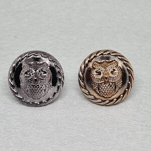 May include: Two decorative metal buttons with owl designs. One button is silver-colored, and the other is gold-colored. Both buttons have a textured owl face in the center and a rope-like border. These buttons are suitable for sewing projects.