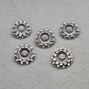 May include: Five silver daisy-shaped embellishments. Each has detailed petals and a central, textured design. The embellishments are arranged on a grey surface, showcasing their intricate floral pattern.