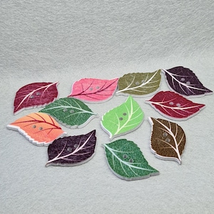 May include: Assortment of ten wooden leaf-shaped buttons in various colours, including green, red, orange, purple, and brown. Each button has two holes and a white background.