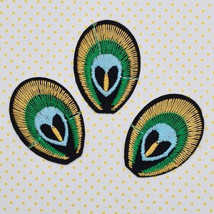 May include: Three embroidered iron-on patches in the shape of a peacock feather with an alien face in the centre. The patches are black, green, yellow, and blue.